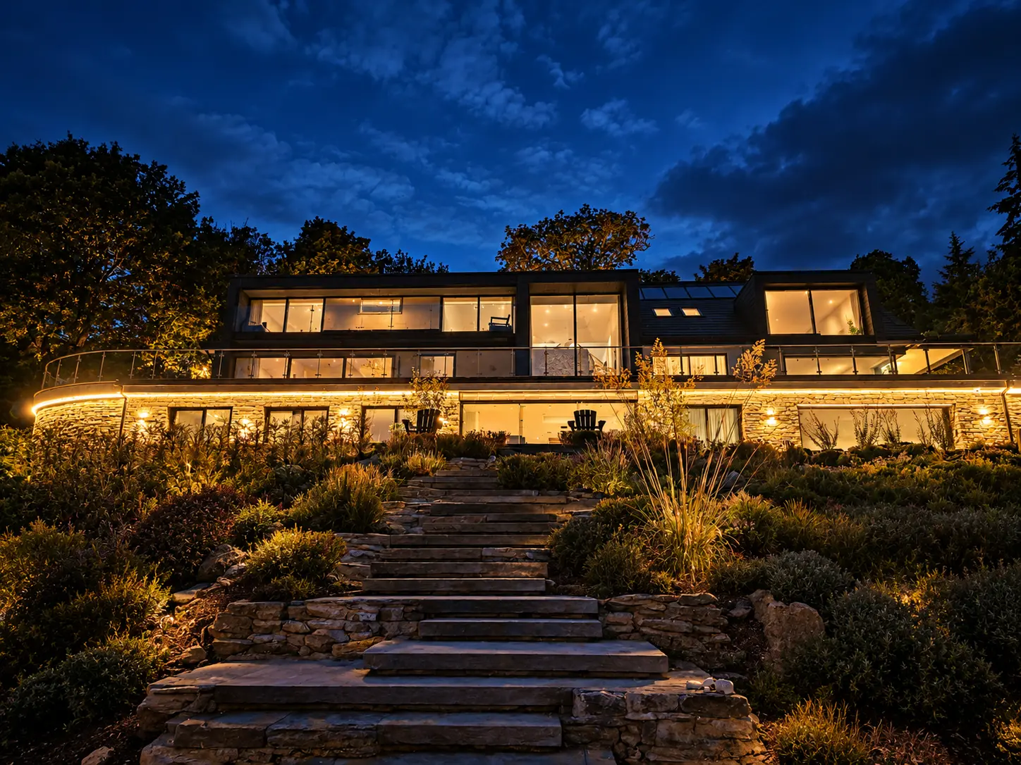 The house lit up at dusk above stone steps and planted terraces