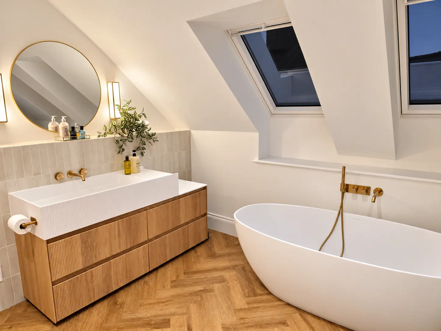 Fluted oak vanity with round brass mirror beside a freestanding bath