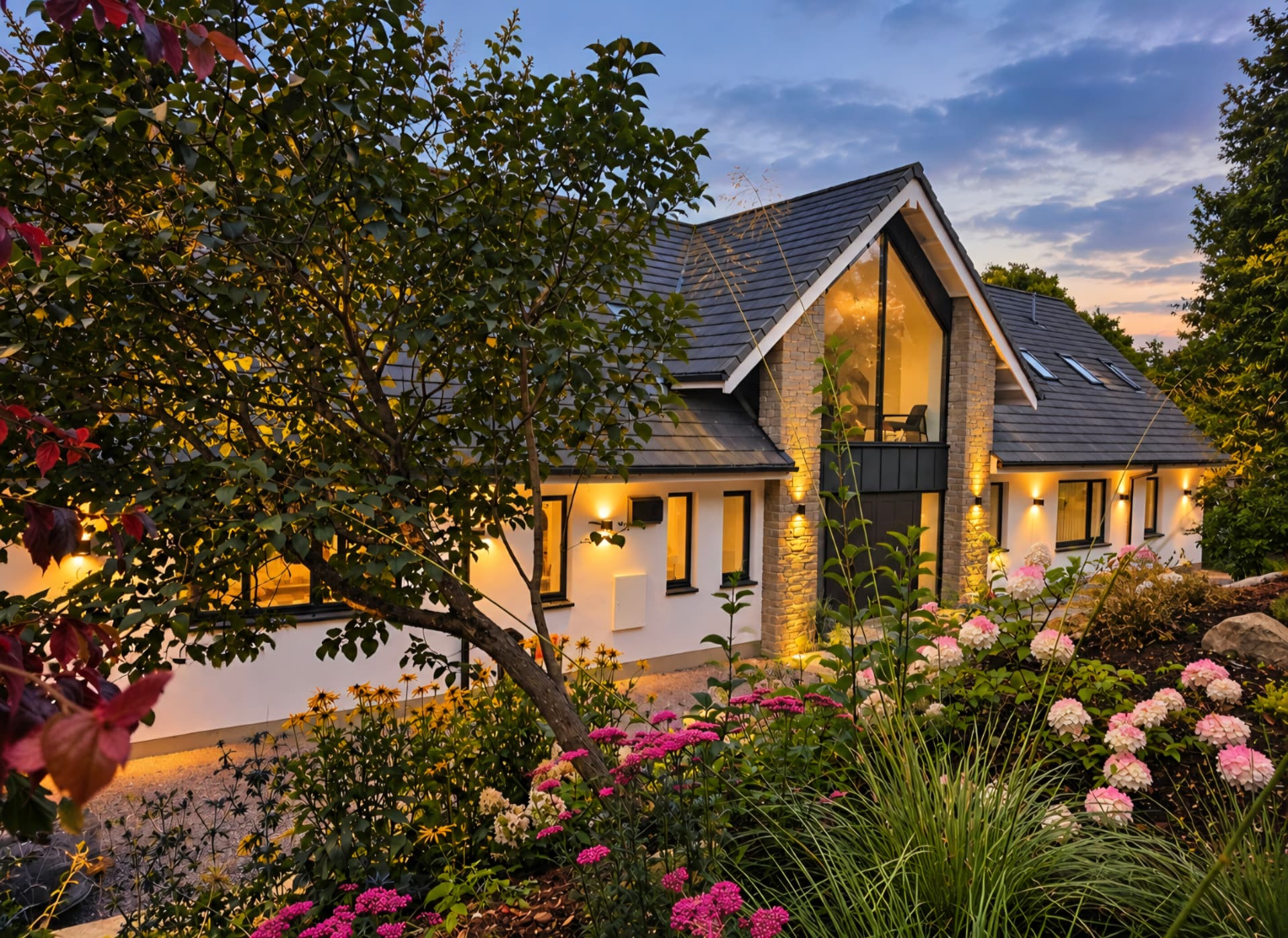 Glazed gable entrance and stone pillars lit at dusk behind flowering hydrangeas